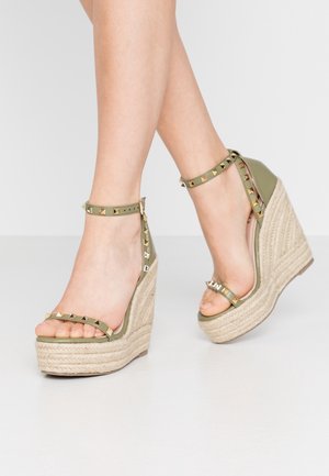 Pies con sandalias de cuña verde oliva con tiras en el tobillo y tachuelas doradas en forma de pirámide, con tacones de plataforma de cuerda tejida.