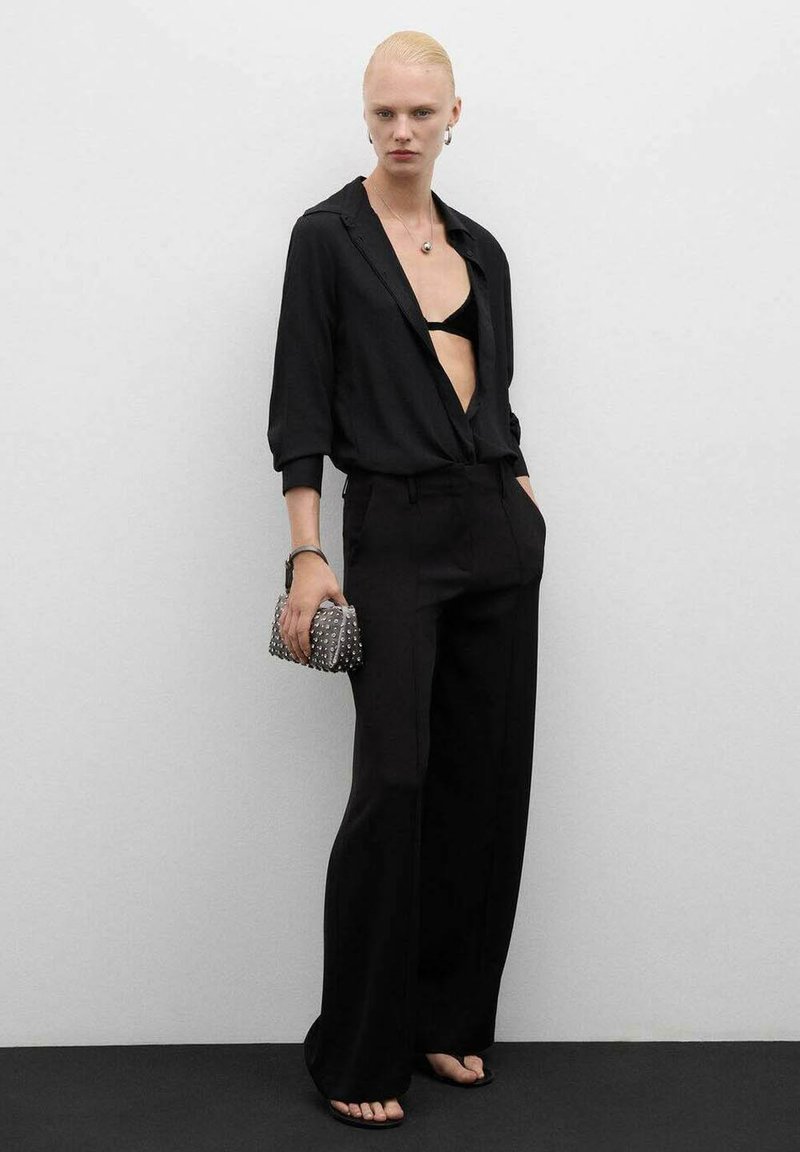 Mujer con pantalones anchos negros y blusa suelta que revela un sujetador negro, sosteniendo un bolso con tachuelas, de pie contra una pared gris lisa.