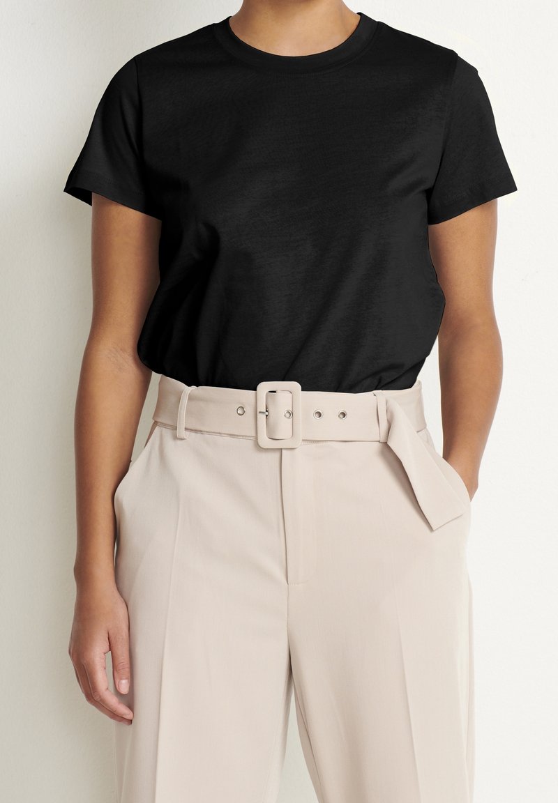 Personne portant un t-shirt noir uni à manches courtes, rentré dans un pantalon beige taille haute avec une ceinture assortie et une boucle rectangulaire.