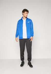 Blå sportsjakke med hvide detaljer over en hvid t-shirt, parret med mørke bukser og sorte sneakers; modelen vender let til siden.
