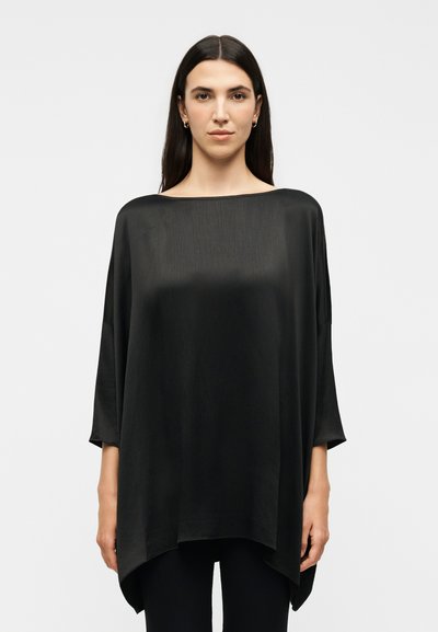 Žena s dugom tamnom kosom, nosi široku crnu satensku bluzu s izrezom do grudi i crne hlače, stoji ispred jednobojne svijetlosive pozadine.