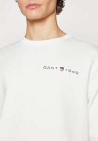 Hvid sweatshirt lavet af blødt stof med rund hals, en lille sort GANT-logo med et skjold og "1949" på venstre bryst.