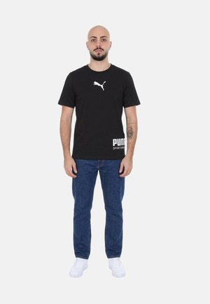 Uomo in piedi che indossa una t-shirt nera Puma, jeans blu, sneakers bianche, con tatuaggio sull'avambraccio sinistro su sfondo semplice.