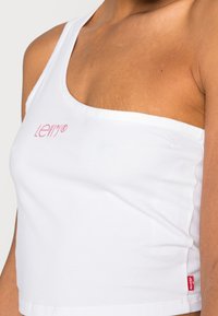 Top corto bianco realizzato in cotone morbido. Presenta una cintura singola e un logo rosa. L'etichetta rossa distintiva sull'orlo aggiunge un tocco di dettaglio.