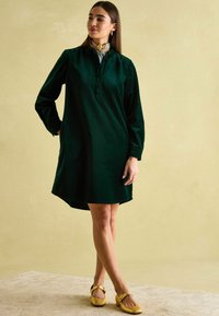 Dunkelgrünes Cordkleid mit langen Ärmeln, Knopfleiste und lässiger Passform, kombiniert mit hellgelben, geschlossenen Schuhen. Glatte Textur.