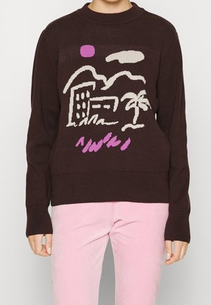 Personne portant un pull marron avec un contour blanc représentant des bâtiments, un palmier, un nuage et un soleil rose, assorti à un pantalon rose clair.