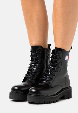 Zwarte veterlaarzen met dikke zolen en zijritsen, gedragen op blote benen, met een Tommy Jeans-logo aan de buitenzijde.