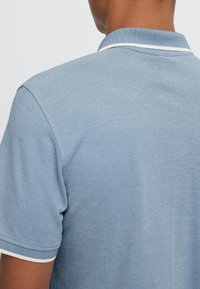 Persoană purtând un tricou polo bleu cu mâneci scurte și bordură albă la guler și mâneci, văzută din spate.