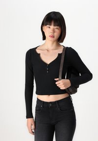 Černý ripovaný crop top s dlouhými rukávy, s hlubokým výstřihem do V a dvěma knoflíky. Model má na sobě tmavé džíny a nese hnědou kabelku přes rameno.