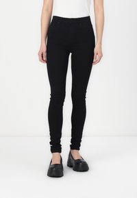 Jeans ajustados de talle alto en negro, hechos de denim, con cinco bolsillos y una textura suave, combinados con zapatos de charol negros y gruesos.