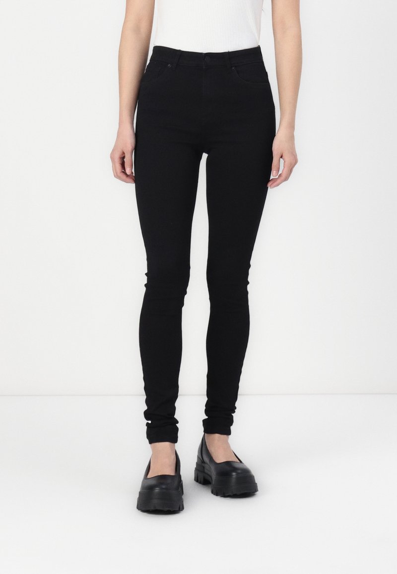 Jeans ajustados de talle alto en negro, hechos de denim, con cinco bolsillos y una textura suave, combinados con zapatos de charol negros y gruesos.