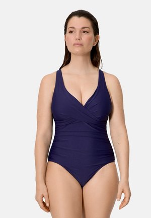 Mujer con un traje de baño de una pieza color púrpura oscuro, con escote en V y detalle de tela recogida, de pie contra un fondo liso y claro.