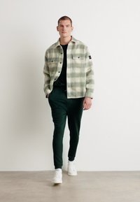 Grønn og grå rutete flanellskjortejakke med knapper foran, to brystlommer, sammen med mørkegrønne bukser og hvite sneakers.
