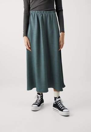 Personne portant une jupe bleu sarcelle mi-longue, un haut noir à manches longues, des chaussettes blanches et des baskets montantes noires, debout sur un fond blanc.