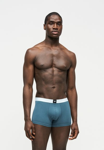 Μυώδης άνδρας που στέκεται σε απλό φόντο, φοράει μπλε εφαρμοστά μποξεράκια με λευκή μέση και ένα μικρό μαύρο λογότυπο.