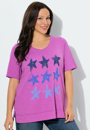 Frau trägt ein lila kurzärmeliges Shirt mit blauen Sternmustern, lächelt, hat lange dunkle Haare und steht vor einem weißen Hintergrund.