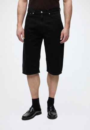 Homme portant un short noir mi-genoux associé à des chaussettes noires et des mocassins noirs brillants, debout devant un fond clair uni.