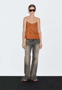 Donna che indossa un top di seta color ruggine, jeans a gamba larga sbiaditi con orli sfrangiati, occhiali da sole scuri e sandali aperti, in piedi contro uno sfondo bianco.