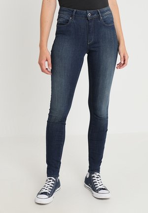 Vaqueros de mezclilla azules ajustados con cintura a media altura, diseño clásico de cinco bolsillos y un ligero desgaste en los muslos. Combinados con zapatillas de lona oscuras.