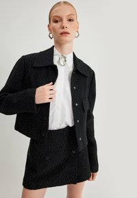 Chaqueta de tweed negra con textura, hilos plateados visibles, diseño corto y cierre de botones, combinada con una falda a juego y una camisa blanca.