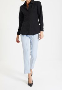 Donna che indossa una camicia nera aderente con bottoni, pantaloni su misura blu chiaro e décolleté neri con punta appuntita, in piedi contro uno sfondo bianco.