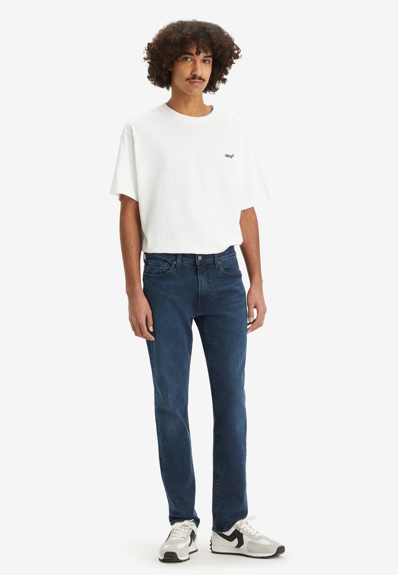 T-shirt en coton blanc avec un petit logo noir Levi's, assortie à un jean en denim bleu foncé et des baskets noir et blanc.