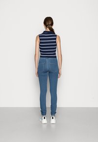 Mujer de pie frente a una pared blanca, vistiendo una camiseta sin mangas de rayas azul marino, jeans ajustados azules y zapatillas blancas.