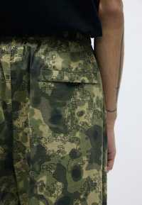 Groene camouflagebroek met een gepixelde patroon, elastische tailleband en een zijzak; gemaakt van een lichte, gestructureerde stof.
