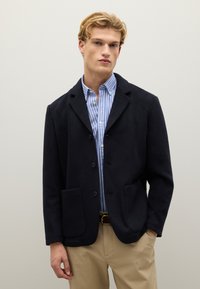 Giovane uomo che indossa un blazer blu navy sopra una camicia a righe blu e bianche, pantaloni beige, in piedi davanti a uno sfondo grigio chiaro semplice.