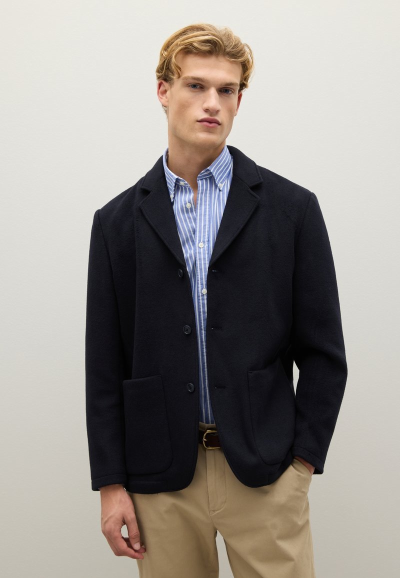 Giovane uomo che indossa un blazer blu navy sopra una camicia a righe blu e bianche, pantaloni beige, in piedi davanti a uno sfondo grigio chiaro semplice.