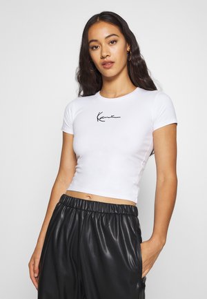 Top court à manches courtes blanc en tissu lisse, avec un logo noir en cursive sur la poitrine ; associé à un pantalon plissé noir.