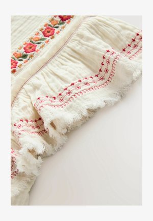 Cremfarbenes Gewebe mit bunten Blumenstickereien am oberen Rand und einem Rüschensaum mit roten gestickten Akzenten und fransigen Kanten.
