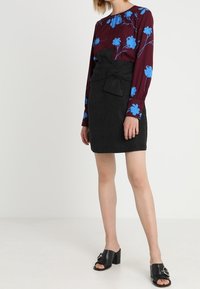 Blouse à manches longues couleur bordeaux avec imprimé floral bleu, associée à une mini-jupe en suède noire ornée d'un détail en forme de nœud, et portant des sandales à talons noires.