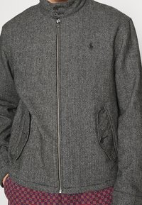 Grå herringbone-jacka med hög krage, framdragkedja och två sidofickor. Har mörka knappar och en klassisk passform.