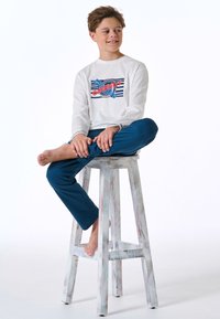 White long-sleeve shirt with graphic design in red and blue. Blue pants. Model seated on a distressed wooden stool, barefoot.