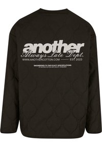 Black quilted sweatshirt with long sleeves, featuring a large white text logo and additional text on the back, smooth texture, and subtle pattern.