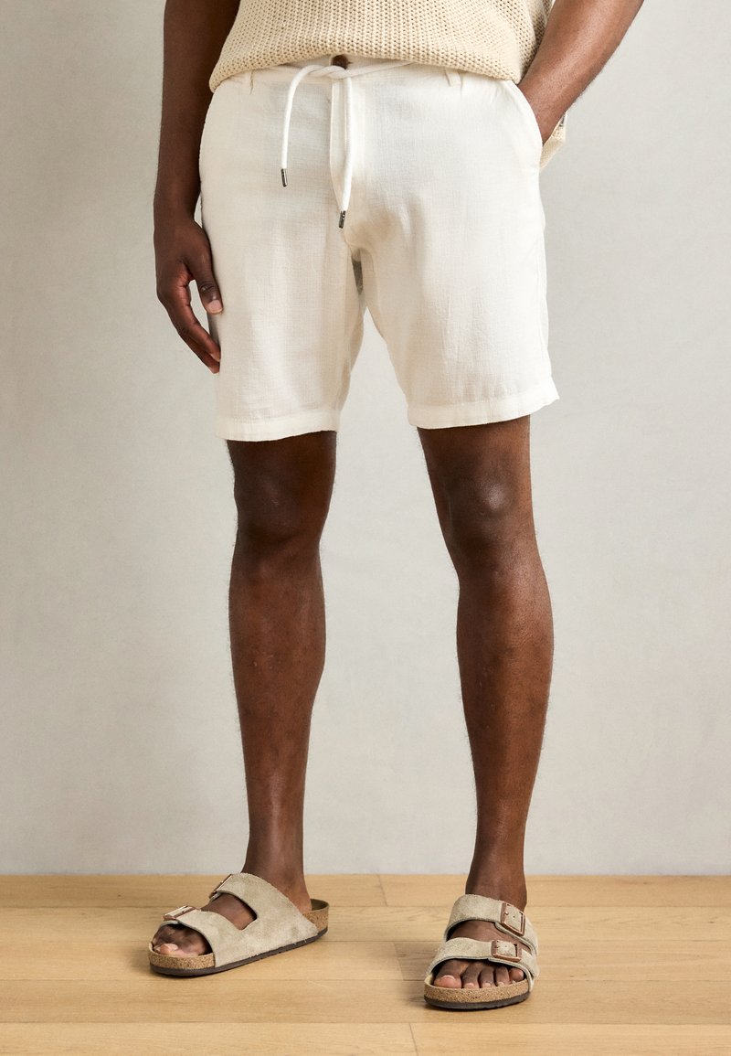 White cotton shorts with drawstring waist, rolled cuffs, and relaxed fit, paired with beige suede sandals featuring adjustable straps.