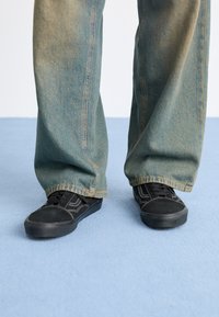 Black sneakers with textured material and white stitching, paired with loose, faded blue denim pants. Background is a light blue surface.