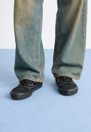 Black sneakers with textured material and white stitching, paired with loose, faded blue denim pants. Background is a light blue surface.