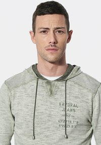 Lichtgroene hoodie met een gestructureerde stof, koordjes met metalen uiteinden en bedrukte tekst aan de voorkant.