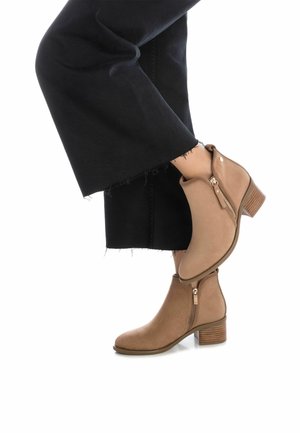 Stivaletti beige con cerniere laterali e tacco in legno impilato, realizzati in materiale simile al camoscio. Indossati con pantaloni neri a gamba larga.