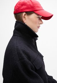 Sort fleecejakke med høj krave og lomme foran, parret med en lys rød kasket. Teksturen er blød, og designet er sporty.