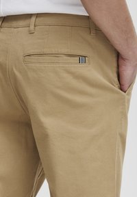 Pantalon en coton beige avec une texture lisse, présentant une poche arrière avec une ouverture horizontale et un détail en accent rayé.