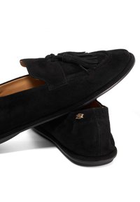 Black suede loafers feature a decorative tassel, smooth interior lining, and a small gold logo accent on the heel. Flat rubber sole.