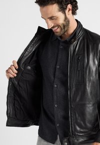 Schwarze Lederjacke mit Reißverschluss, Innentasche und braunem Kragen. Getragen über einem dunklen Hemd mit Knöpfen. Glatte Textur und taillierter Schnitt.