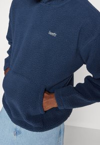 Maglione in pile blu navy con superficie strutturata, tasca a marsupio e logo Levi's bianco sul petto. Vestibilità casual con polsini a coste.