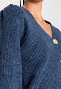 Cardigan en tricot bleu foncé, avec un col en V, des manches bouffantes et deux grands boutons de couleur claire. Tissu texturé.