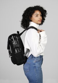 Sac à dos noir avec une finition mate et des accents brillants, présentant des rayures verticales et des détails de fermeture éclair. Porté sur l'épaule avec des bretelles rembourrées.