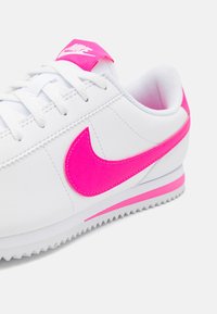 Fehér sneaker rózsaszín akcentussal. Tartalmaz egy rózsaszín swoosh logót, rózsaszín sarokpántot, és texturált fehér felsőrészt. Fűzős dizájn, gumitalppal.