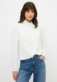 Blusa bianca a maniche lunghe con colletto alto e pieghe sottili, abbinata a jeans in denim blu con decorazioni simili a perle.